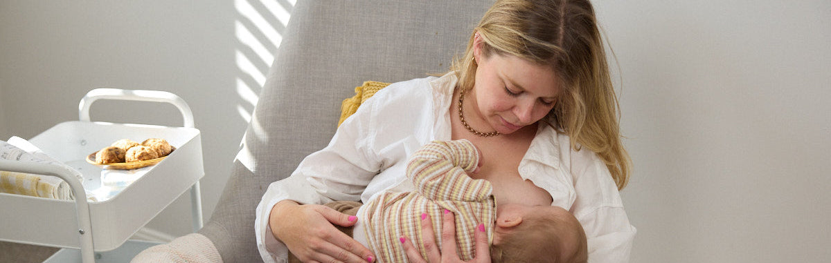 Woman breastfeeding a baby in a chair with a neutral background and plate of lactation biscuits