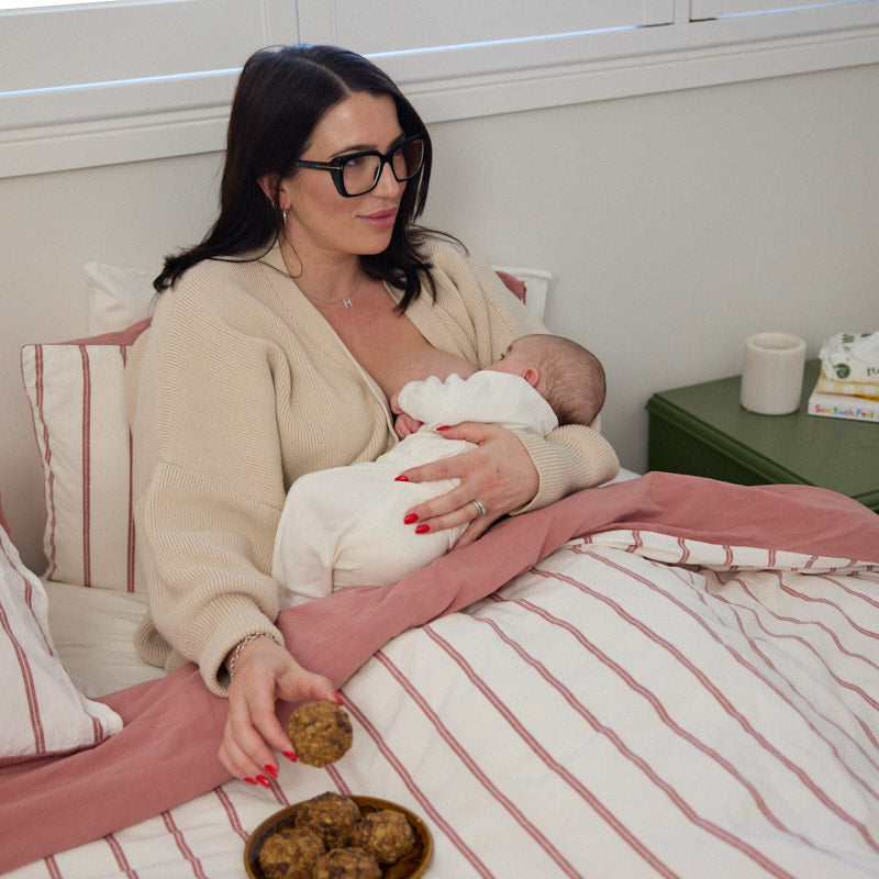 Woman breastfeeding a baby in bed with a plate of breastfeeding biscuits.
