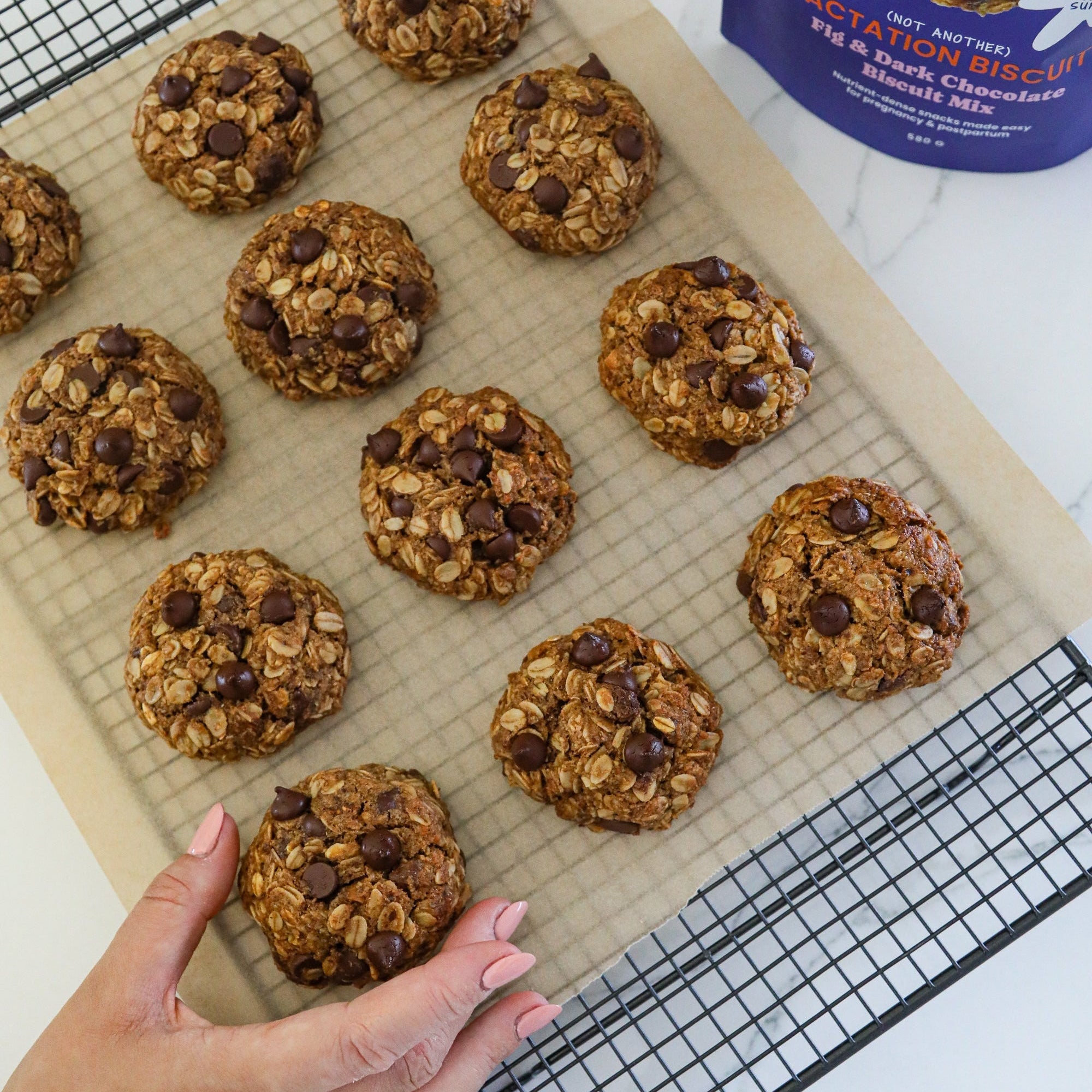 Fig & choc lactation cookies on a cooling rack with a hand reaching out, next to a package of baking mix.