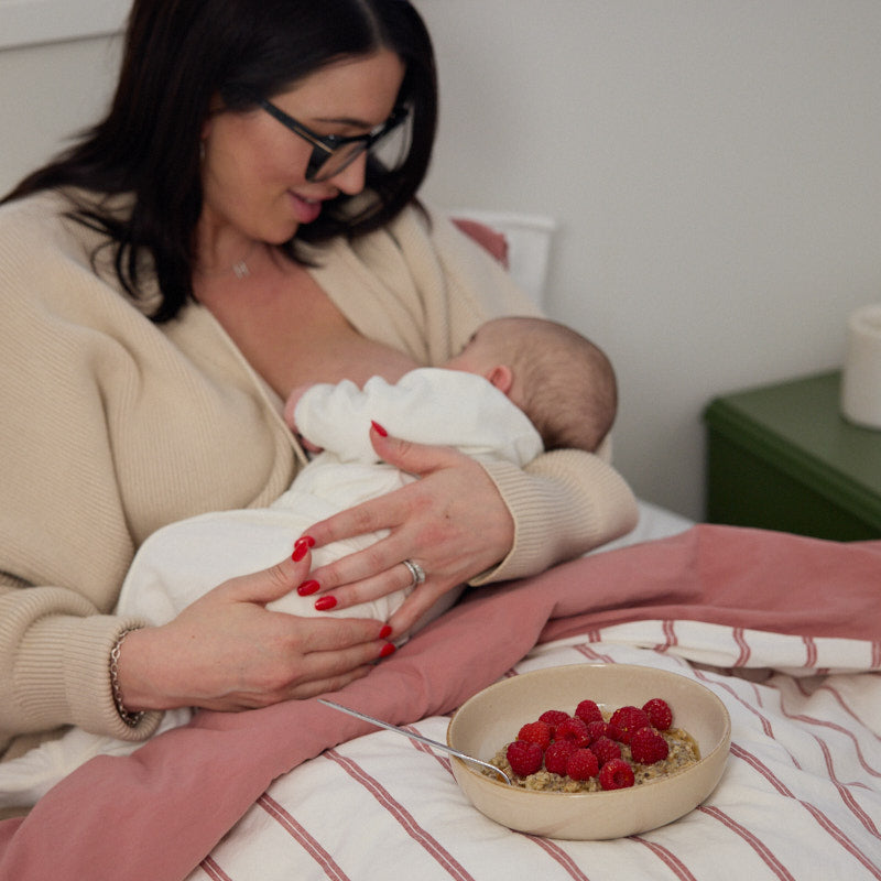 Mum breastfeeding newborn in bed with a bowl of raspberry-topped postpartum food beside her, nourishing support for early motherhood.