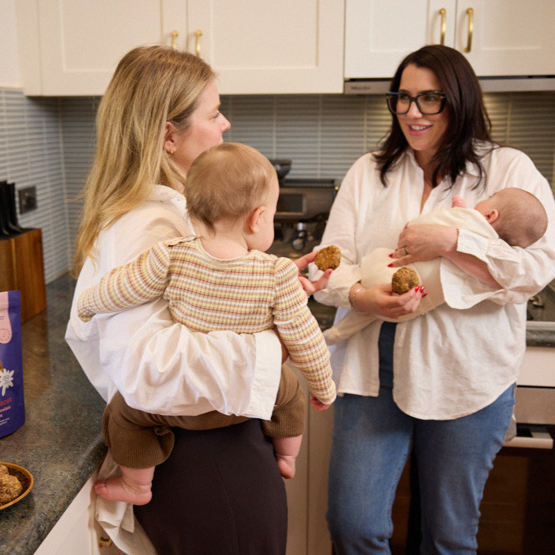 Two mums chatting in the kitchen with babies in arms, sharing Nibbles & Nurture lactation biscuits